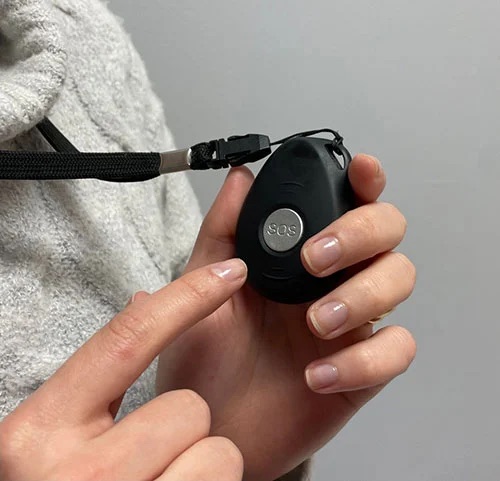 The GPS pendant held in a hand, showing its SOS button.