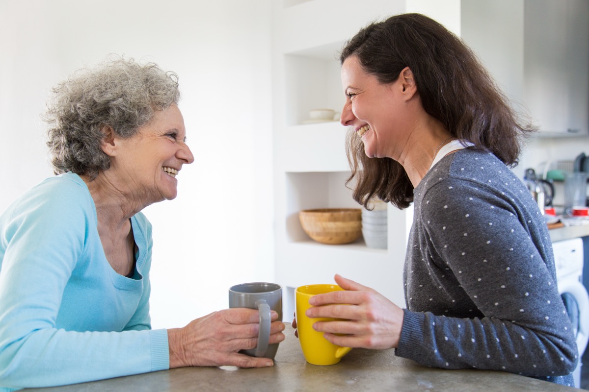 An adult daughter laughing with her mother over coffee in the kitchen.